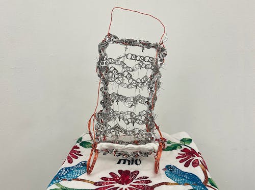 A cart sculpture made out of soda can tabs. The body of the structure is silver and the handles and wheels are made out of orange wire. In between the front wheels, there is air-dry clay. Small silver crosses are all over and between the cart. The cart rests on top of an embroidered pillowcase with two birds and has writing that says “Amor mio.”