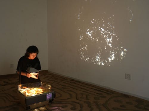 Image of person kneeling by an overhead projector, the floor is covered in sand thats arranged to form a circuit board, orange peel sand covers the top of the overhead and is reflected onto the wall on the left of performer who holds a piece of yellow lined paper. 