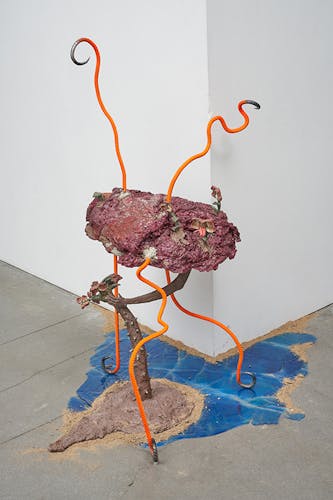 There is a blue pool of resin that resembles water on the ground, it has sand around its parameter. There is ceramic crown thorn orchid growing out of a brown concrete mound. It's top is leaning to the left. There is a maroon leather body with exploding maroon foam held up by three pieces of neon orange rebar with curled legs. Coming out of the top of this leather/foam body are two neon orange curled rebar horns. There are several crown thorn orchid flowers coming out of the leather/foam mount and one flower with a tongue curled. From underneath the leather/foam mound an arm reaches out to try and grab the crown thorn orchid on the ground as it leans away.