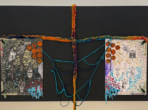 The piece "Deeper Than Ink," is a large installation onto a black wall. There are two panels that are separated by thick, crocheted, rainbow yarn that forms a draping cross. The panels are covered in inked paper towels, tongue depressors, tattooed oranges, and other supplies collected during the tattoo process. There are also drawings of church bells, a triumphant male body, a machine tattooing hand, and a stick and poke tattooing hand. 