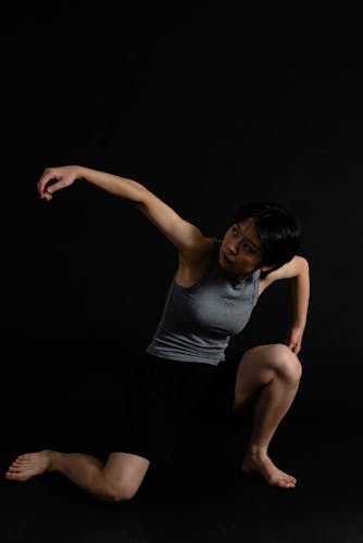 In the dark background, there is a dancer right in the middle of the picture. She is doing the posture of a very dynamic movement. Her pelvis is really low to the ground, and she is with right knee down on the floor, the other left knee is up. The upper body is slightly leaning into the front with her right arm raised higher than shoulder and is slightly bent in the air and her right arm does the same movement but bending more to 90 degree. At the end, her head is looking towards right arm. 