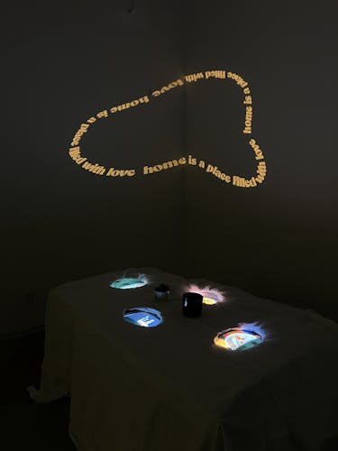 The installation involves a surface with a white cloth draped over and four holes cut out. Beneath the surface are four small old TVs, with three of them playing videos of persons' hands eating a meal, and the other one playing a clip from a cartoon explaining where food goes after eating. These videos play through the four holes that are cut out. On the walls behind the table is a projection of a phrase "home is a place filled with love" looped into an organic shape and rotating. 