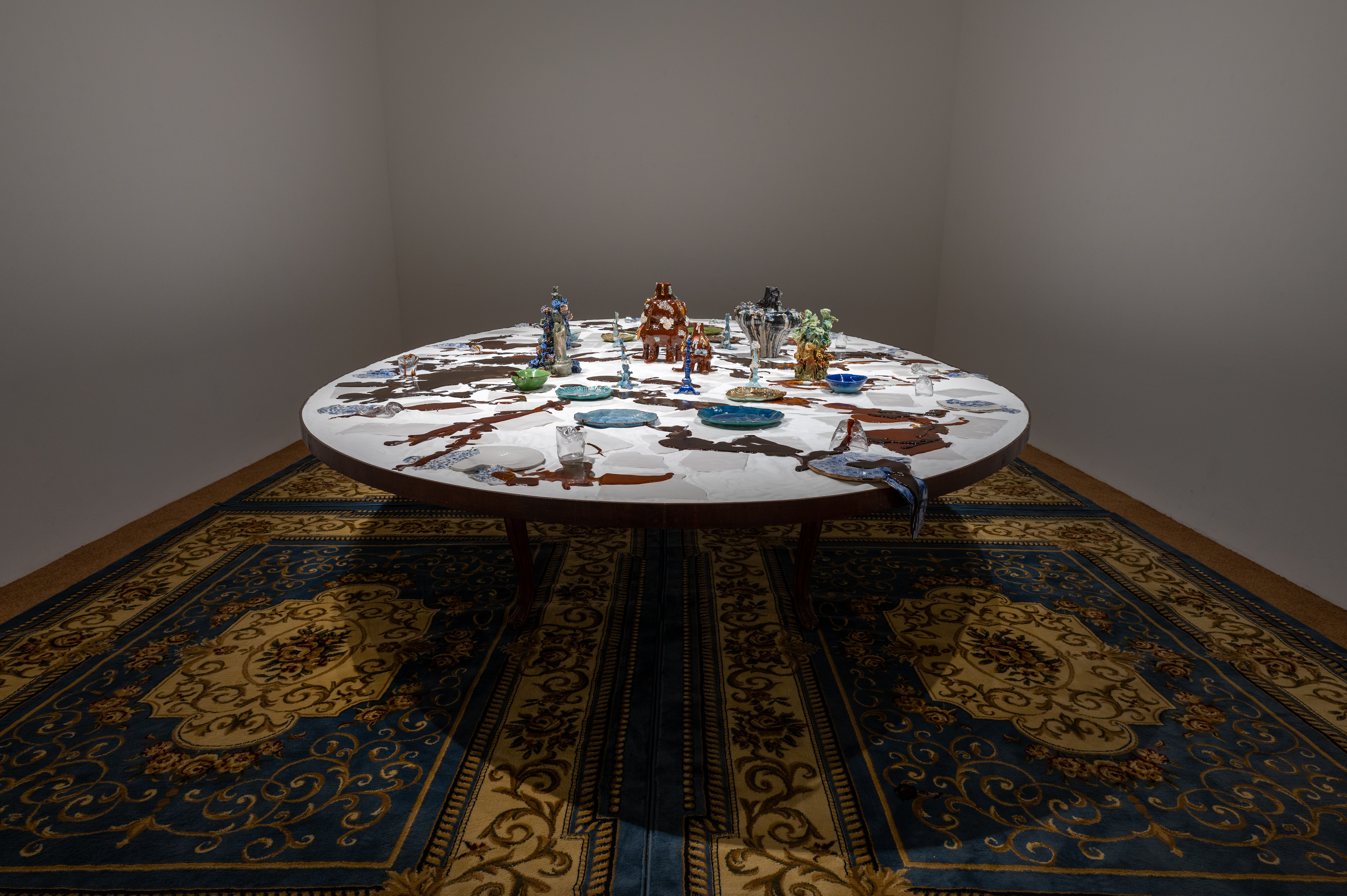 Image of a large circular table with ceramics and melting blue and white porcelain. Candies brown sugar is poured on top of the table creating pools of brown. 