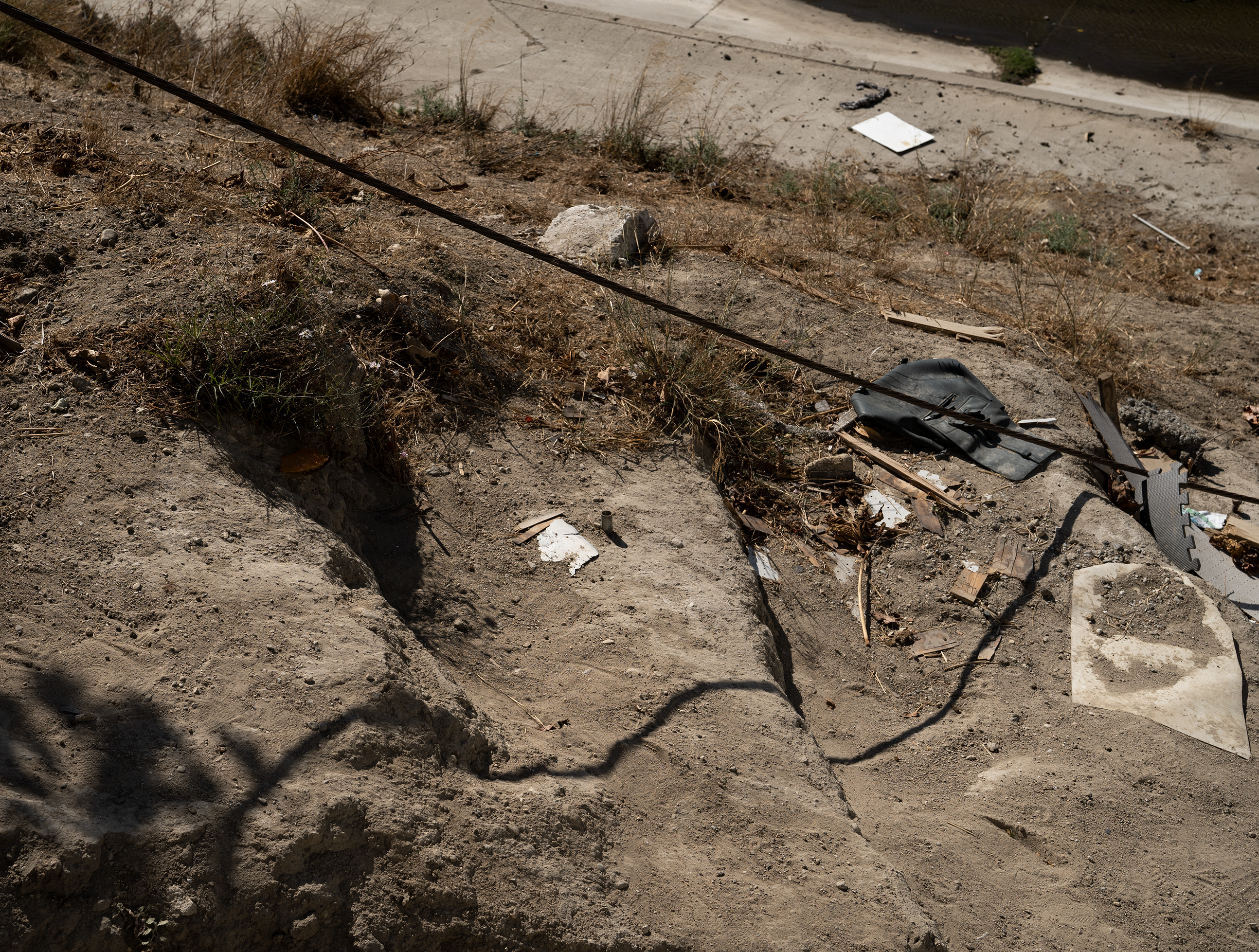 This is an image captured by the banks of the river next to Baldwin Hills, Culver City. It points towards the debris: sand, dirt, trash, grass, etc. The shadow of a electric chord passes through all these objects and forms a line across the image.