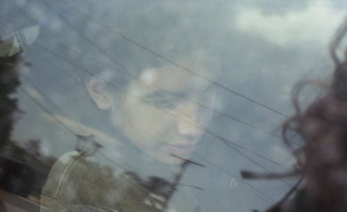 A photograph of a young androgynous person's side profile through a window. They are visible through a reflection of power lines and a person's hair in the bottom right corner.