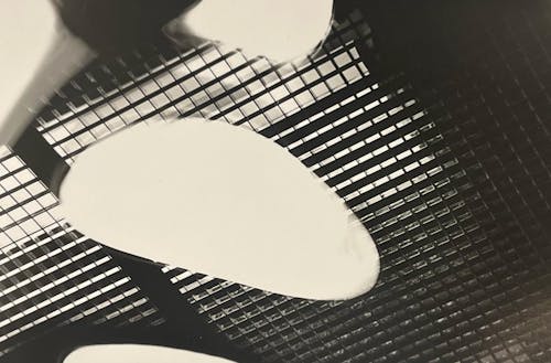 "Reminisce," a darkroom print, features an abstract composition with a strong contrast between light and dark areas. The image captures a grid-like pattern in the foreground, with soft, irregularly shaped light patches scattered across it. The interplay of shadows and light creates a sense of depth and texture, evoking a nostalgic and contemplative mood.