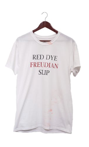 White t-shirt hanging from brown hanger against white background with "red dye freudian slip" printed in the center of the shirt. The words red, dye, and slip are printed in black and freudian is printed in red. There are red Cheeto dust stains on the collar and bottom right of the shirt.