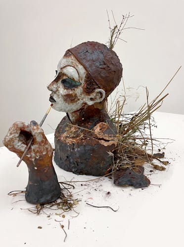 Life-sized ceramic bust of a drag performer wearing a wig cap and face of makeup. A hand holding a paintbrush accompanies the figure. The back of the bust of cracked open and filled with Los Angeles weeds and native plants. The makeup and surface of the piece is painted largely with iron oxide pigments in a variety of colors, crumbling from fired clay to powdered form from the open back, referencing decomposition. 