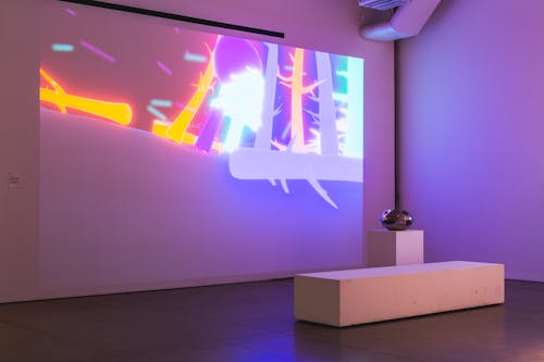 Projection of brightly coloured 3D trees in various orientations on a terrain, mostly purple, blue and orange. In front of the projection on a pedestal stands a handpan drum shaped controller for the projected game. A white bench stands in front of the installation.