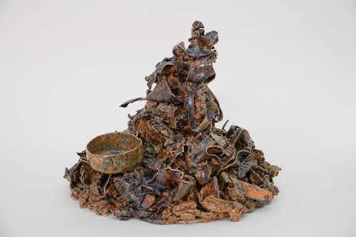 A ceramic sculpture of trash, glazed with non-traditional materials on a white backdrop. The color of the glaze is a splotchy combination of rusty orange and metallic black/gray. Some forms are more recognizable than others in the pile, such as the handle of a teaspoon, and a cup measurement. 