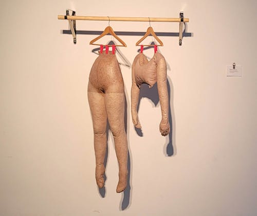 a sculpture of legs, panty hoes filled with wood shavings and stiffened with glue, and a torso, similarly made, are hung by a wooden coat hangers against a white wall. The sculpture is resting on a wooden dowel silver brackets that mimic a closet.