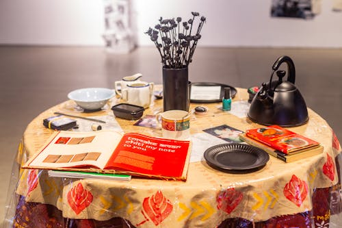 Round table with various items on it. A hand printed textile tablecloth is laid upon it. Some items like the flower vase, plates, and teapot are spray painted black. A printed book is at the front of the table. Other items include pictures of the artist and her grandmother, handwritten recipes, utensils, coffee mugs and a recipe book. 