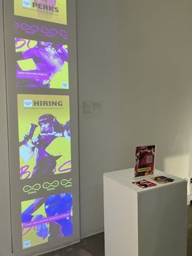 This picture features a projection scantily-dressed video game women are featured. There are four graphics featuring these women vertically on the wall, each one with a text about hiring women for Diversity, Equity, and Inclusion efforts. Next to the projection is a pedestal, with a hiring ad sitting on top of it. It has a QR code leading to a website.   