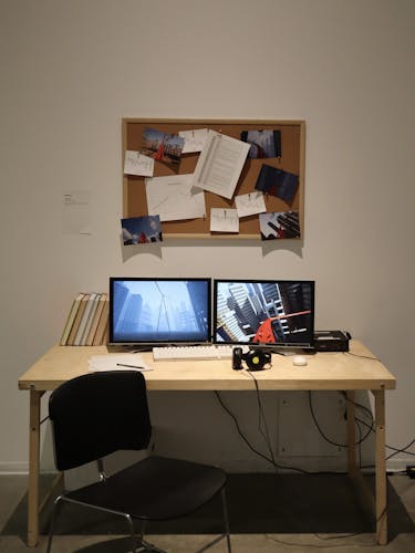 A gallery set up with a wooden desk, black chair, dual monitors showing a roller coaster with cityscapes, a keyboard, mouse, headphones, and books. A corkboard above displays photos, graphs, and documents.