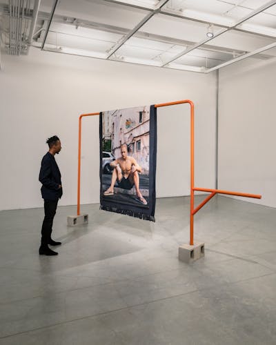 A man sitting in front of a sculpture in a gallery. The sculpture is composed of a bright orange pipe structure with a computer-generated image printed on a textile that hangs over it. The printed image depicts an eastern-european man sitting on the side of a road, looking at the viewer.