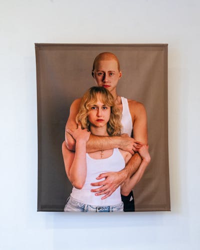 A textile print hanging on a gallery wall. The print depicts a man hugging a woman, they both have the same facial features.