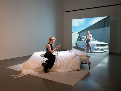 An installation of a bed, a grey sweep and a projection in a gallery. A woman sitting on the bed, looking at a phone. In the projection, a man is looking at his phone, leaning on a 3D representation of a car, located in an industrial setting. 