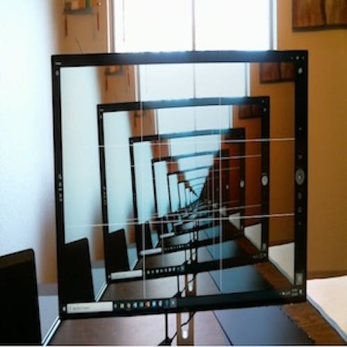 A monitor is placed on a dark-brown wooden desk with a window and some artwork on the wall in the background. This appears to be a distorted image of the monitor which multiples its original image by manifolds.