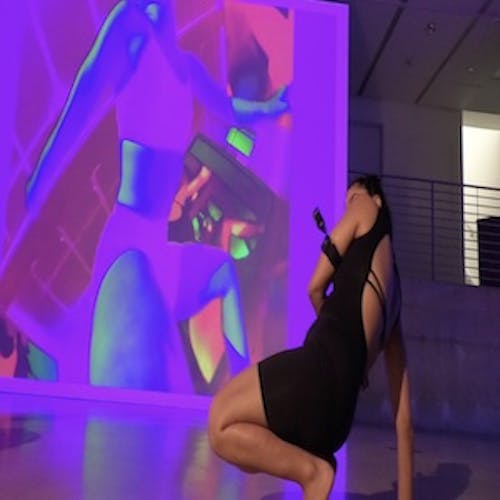 Indian dancer, wearing all-black leotard and shorts, squats low to the ground, balancing herself on her feet and one hand. Her knees are bent, and hips make contact with the heels. Her other arm maps the lower half of her body with the go pro camera attached to her upper arm. In the background, we see an image of another dancer’s body projected on a wall. She is wearing a black sports bra and shorts; however, her image appears to be fluorescent pink in color with the use of a filter.