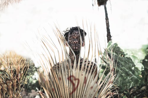 A black man with bare chest in face paint covered by a large palm frond with a red "B' on it