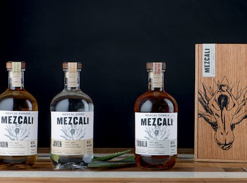 A photograph of three glass mezcal bottles arranged in a row to the left and its wooden box packaging on the far right against a black backdrop and sitting on a wooden table with a few agave leaves next to the center bottle. The image is showcasing the branding of the company which is comprised of a stenciled black illustration on a white background of a woman facing away and kneeling while holding a cup above her head with a big agave plant placed in front of her.  The brands name Mezcali rests above the illustration in bold uppercase black letters and each bottle has a different type of mezcal neatly arranged in a paper sub ribbon that is wrapped around the bottom of each bottle. From left to right the ribbon reads espadin, joven, and tobala with illegible small text to the right of each.