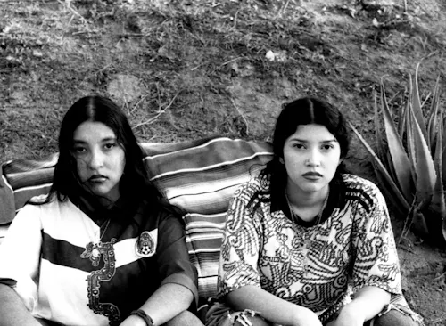 A black-and-white photograph featuring two young women with long, dark hair sitting on a blanket outdoors, against a dirt hillside backdrop. The women wear patterned soccer jerseys—one with a large dragon design and the word "MEXICO" on the chest, the other with an abstract pattern and "MEXICO" written across the front. Both women look directly at the camera with neutral, serious expressions. An aloe plant is visible in the background beside one of the women. The setting includes a striped blanket behind them, and the overall atmosphere evokes a sense of cultural pride and quiet resilience.