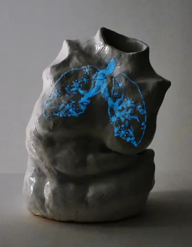 In a dark room is a white ceramic sculpture of a female torso slumping slightly to the right. The torso is stylistically similar to Roman marble sculptures, having no arms or head. On its chest, a drawing of lungs with small cartoonish virus motifs glows blue, reminiscent of an MRI.