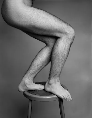 A black and white studio portrait of two hairy legs with lighter skin, balancing upon a metal stool. The leg closest to the camera is stepped slightly in front of the back leg, concealing the genitalia of the subject. The background is a dark gray.