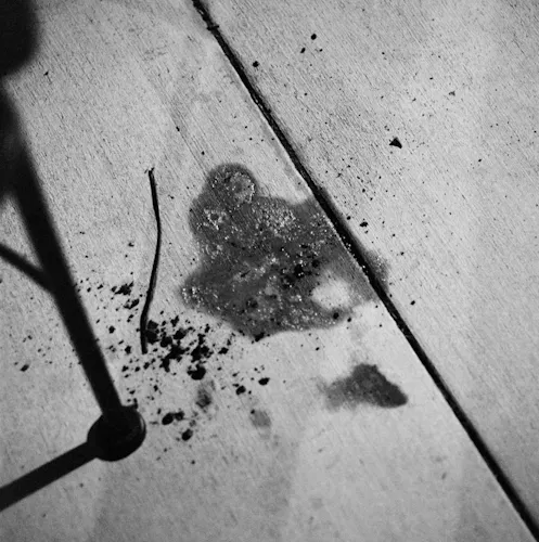 An ambiguous puddle of what appears to be spit is pooled on the concrete. A metal chair's leg encroaches slightly into the frame, and the faint shadow of the photographer's legs is cast over the puddle.
