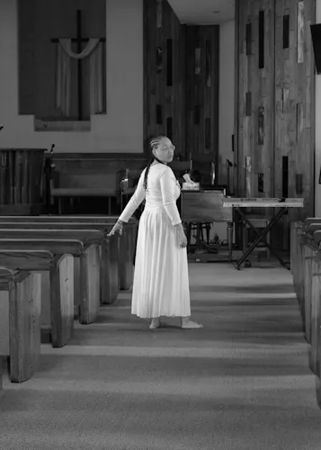 a black and white photo of a women in a white dress standing in the middle of an aisle in an empty church while touching a pew and looking back at the camera with a large cross in the background