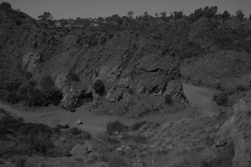 A camera looks down from a hill onto a canyon made by a former mine. It now looks like it had always been there. The print is dark, as if it is night, and there are a few spiraling stone labrynths in the distance, with homes just on the horizon.