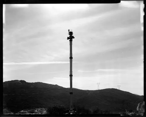 A black and white contact print which includes the black film edges, some light leaks, and chemical stains. In the center of the image is an autonomous surveillance tower, built by Anduril Industries for the "virtual border." Below, in the far background, are scattered buildings and roads and along the horizon is the faint silhouette of the built border wall.
