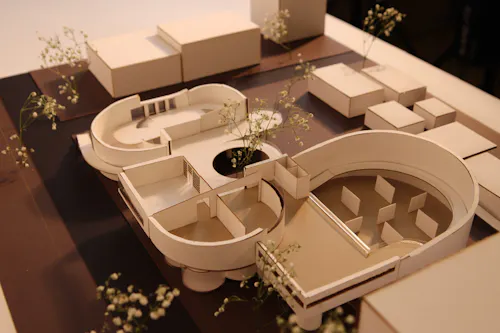 A model of "The Common Ground" sits on a base that depicts the surrounding site, including streets and neighboring buildings. The building has many curves, resembling that of Le Corbusier's Villa Savoye. The model's interior and program are shown. This includes gallery spaces, art studios, and open spaces. Materials used to build this model include museum board, bristol, cardboard, vellum, and baby's breath flowers. 