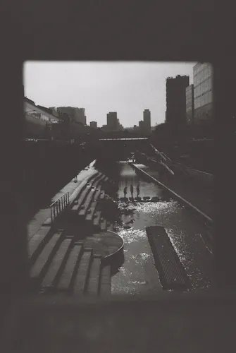A black & white photo shot on film, depicting a city skyline and figures going across a body of water on stone steps.
