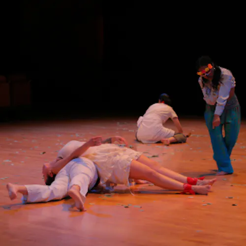 In the foreground, a Latinx performer with dark hair, a foot and forearm tattoo wearing an off white sleeveless top, ruffled shorts and a red bandana anklet on each foot, rolls across the back of another light skinned Latinx performer dressed in all white in the foreground. To the right, a Black performer wearing peacock blue-colored pants and a silky light blue button up with the mid section exposed, a Día de Muertos skeleton half mask crowned with pink, orange and yellow flowers, hovers over the two dancers in the foreground. In the background, a performer dressed in a white top and dark green pants layered with a white waist wrap spreads Eucalyptus leaves across a "blonde" wooden stage floor. .