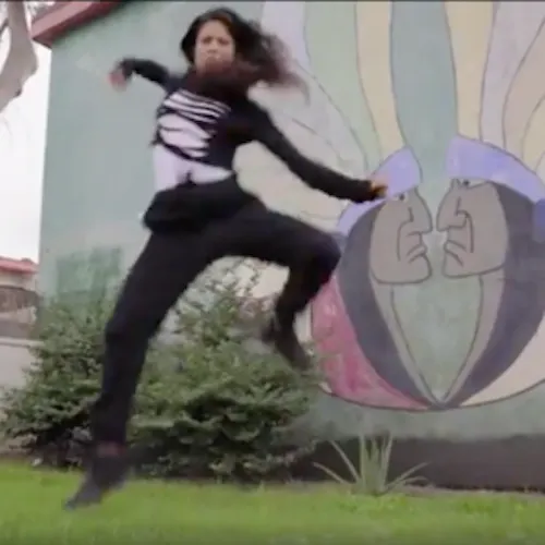 In an outdoor area with a mural of two Indigenous American men facing nose to nose, wearing yellow headdresses in the background while a Latinx performer  wearing black pants, a ripped black hoodie and jumps in the air with knees bent and arms flexed out wide.