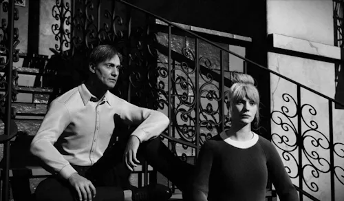 A black-and-white still frame recreation from La Jetée, showing a man and a woman seated on a wrought-iron staircase. Harsh lighting casts deep shadows across their faces and the ornate railings behind them. The man gazes ahead thoughtfully, while the woman stares into the distance, evoking the film’s haunting, time-suspended mood.