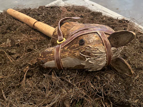 A ceramic horse head (from the neck up), its skin is a muddy spotted brown with a curved white stripe that runs down the center of the horse's face to its nose. The horse's mane reaches just above it's dime size black eyes and it is white with hits of brown in-between the strands. Wrapped around the horse's face is a warm brown halter. Stitch-lines outline the horse head's most prominent features––the nose, the neck, the edges of the halter. Just below the neck of the horse is its body; which is a golden brown ceramic wooden stick with warm red undertones––wood grain covers the entire surface of the stick which is broken at the center. These ceramic pieces lay on a large pile of gardening waste material (e.g., dried leaves, dried pine tree needles, soil, grass, and roots) that is a dark brown shade with hints of light brown scattered within it. 