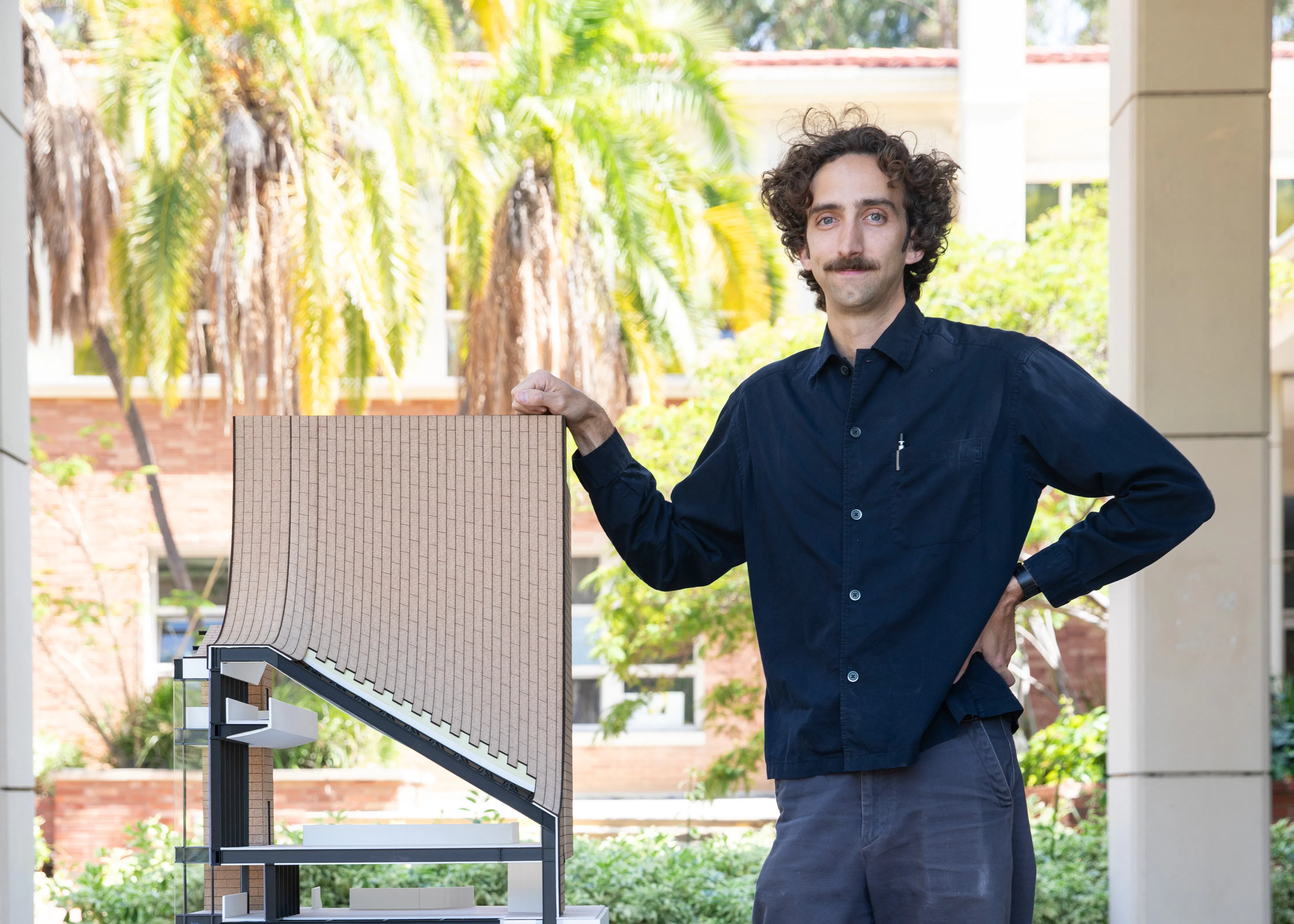 Hunter Blackwell stands in the Perloff Hall courtyard beside a detailed architectural scale model featuring a dramatic curved facade with a textured grid surface. He has curly dark hair and a mustache and is wearing a navy button-down shirt and gray pants, with one hand resting on the model