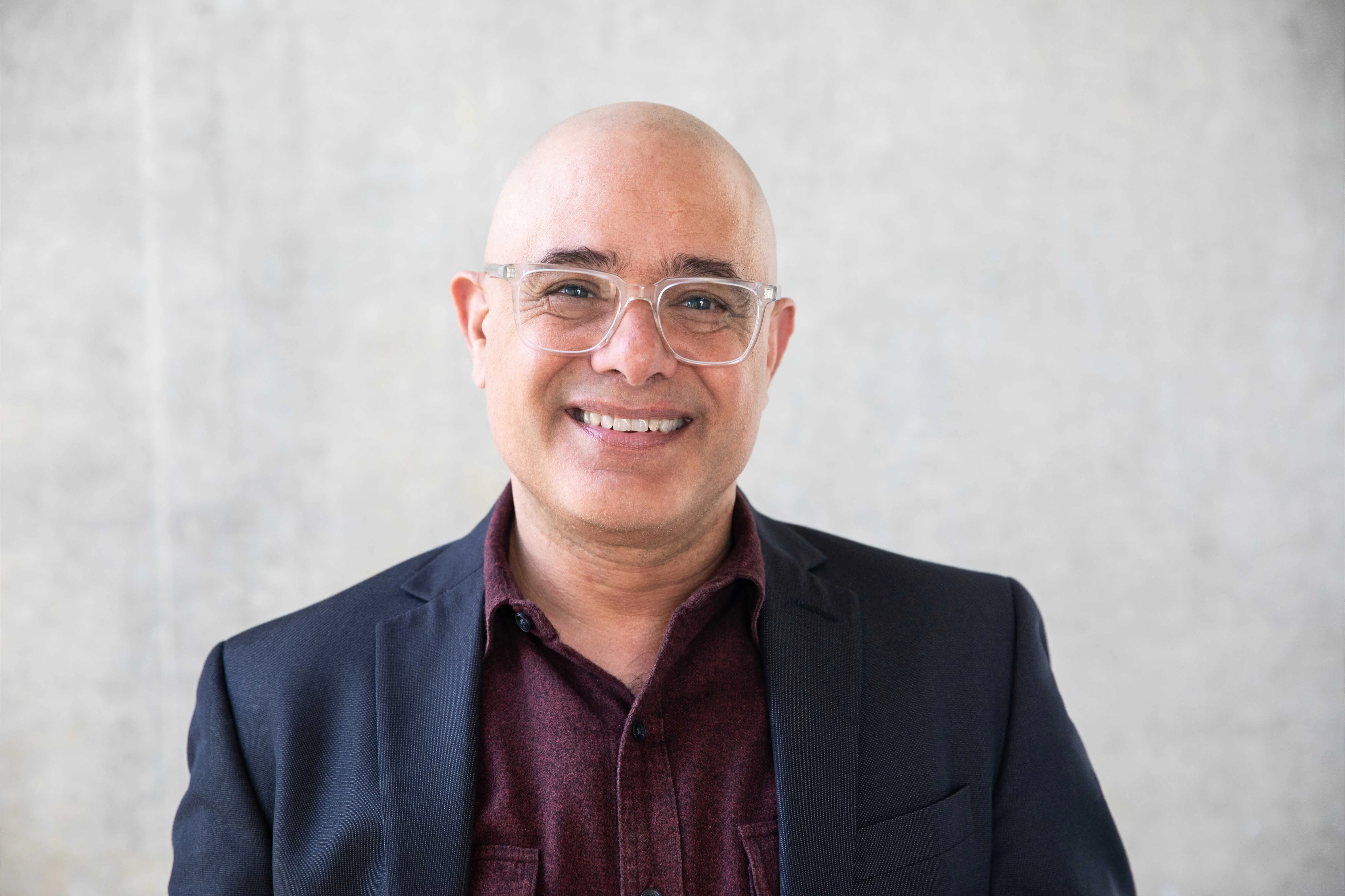 Headshot of Dean Lionel Popkin smiling warmly at the camera. He is bald and wears clear-framed rectangular glasses, a burgundy button-down shirt, and a navy blazer. The background is a neutral light gray textured wall.