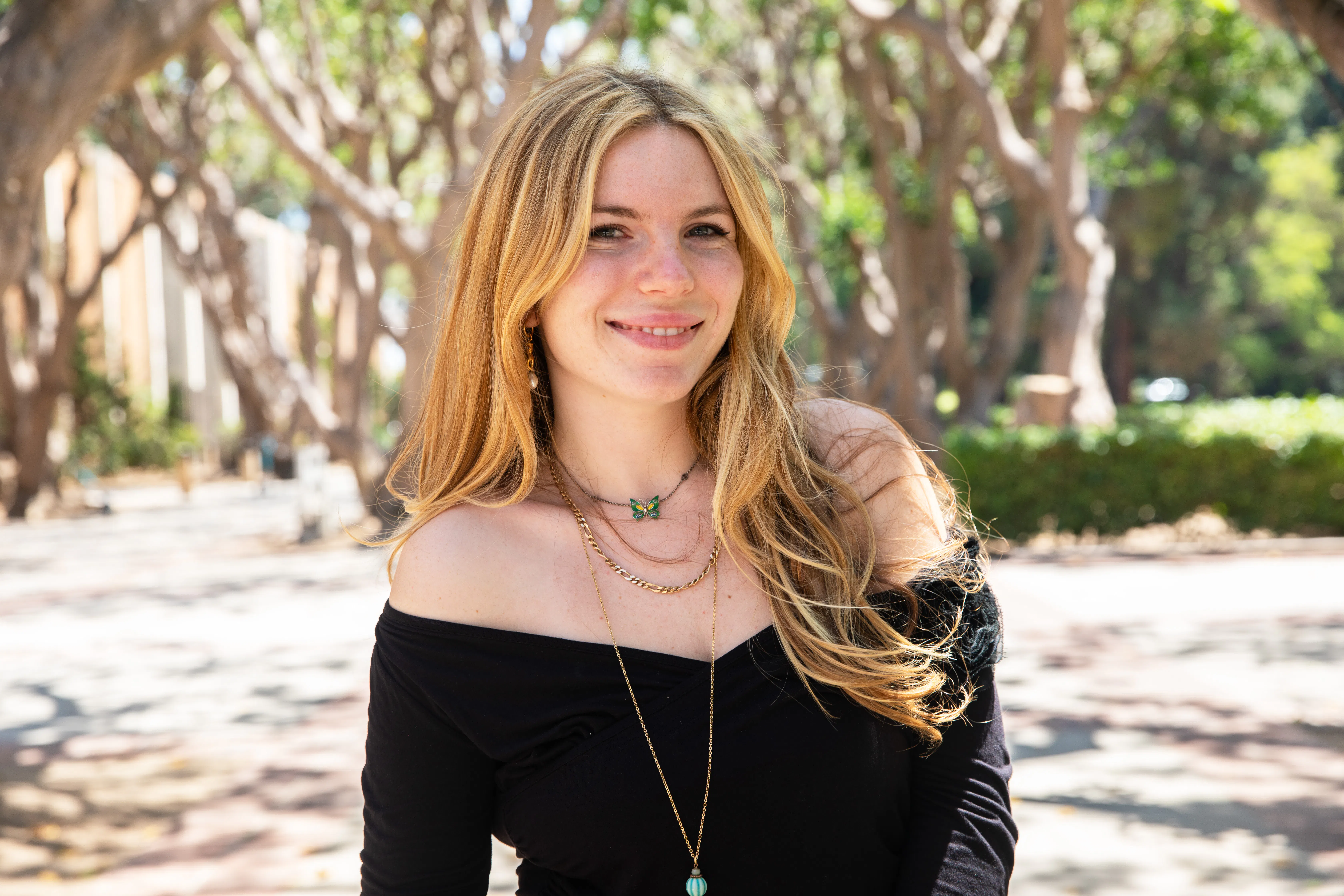 Headshot of Sarah Lustgarten, undergraduate student speaker, smiling outdoors on campus. She has long blonde hair and wears a black off-shoulder top with layered gold necklaces including a green butterfly pendant. A sunlit tree-lined walkway is visible in the background.