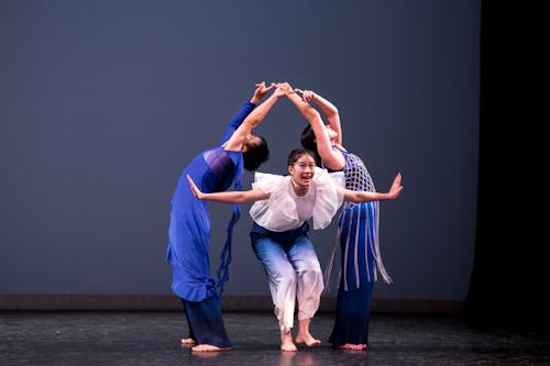 Three dancers engage in child's play onstage, wearing a color palette of deep blues, white, and silver, in various textures that bring a sense of lightness and flow to their physicality. Two of the dancers, one of Chinese ancestry and the other of Filipino ancestry, are leaning backwards and are conjoined by their fingers. The third dancer emerges between the middle space created by the other two. Also of Chinese ancestry, this dancer holds their arms with palms flexed, and has an expressive look of curiosity and excitement, as if making an announcement to the audience. Photo by Raphael Arndt features Lingfeng Yi, Bianca Yen, and Mikayla Knestrick.