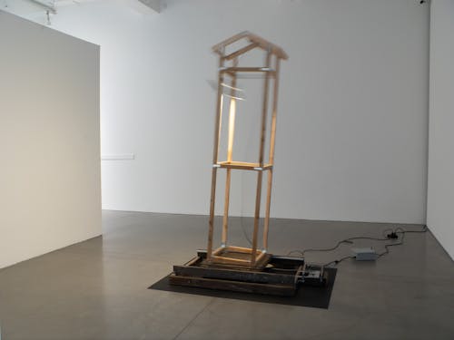 A wooden sculpture installed inside a gallery space. The sculpture consists of a wooden base attached to a wooden gantry on wheels, with a structure that resembles the framework for a house on top. A pendant lamp hangs from the sculpture, illuminating it and the space around it.