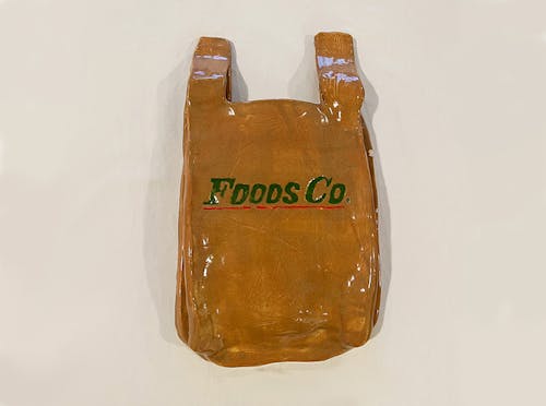A brown plastic grocery bag from “Foods Co” that is made out of ceramics. In the middle of the bag, it says “Foods Co” in green blocked letters and is underlined in red.