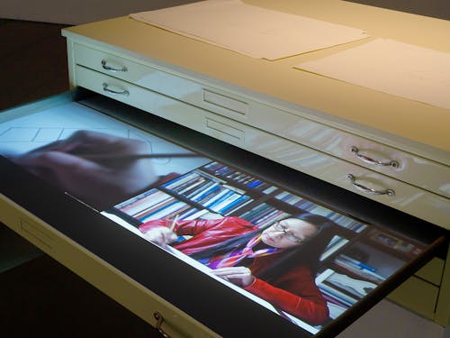 A steel flat file painted with cream automotive paint has a drawer opened. Inside the drawer plays a split-screen video projection. On the left channel, an older Chinese woman sits with her back turned inside a car. On the right channel is a close up of a hand holding a pencil as it draws on a piece of paper.