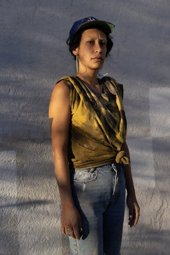 The photograph is vertical. A woman is standing in front of a stucco wall. She is wearing a blue LA cap, hoop earrings, and an earth yellow tie dye shirt. The light hitting her is golden, like her shirt. She stares directly at the viewer. 