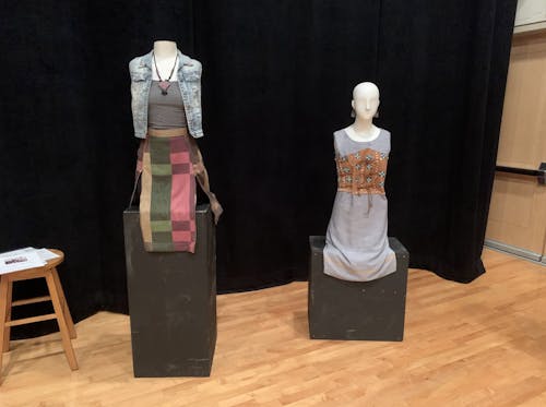 Two half body mannequins rest side-by-side on separate podiums. The leftmost mannequin wears a blue denim vest, grey camisole, black necklace, and checkered green, purple, and beige skirt. The rightmlost mannequin wears silver chandelier earrings and a grey shift dress and a brown corset with white shells sewn to it.