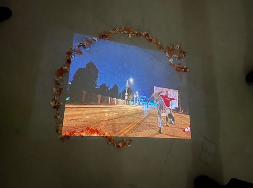 Image still of video installation on the floor, aerial view of dried orange peel cut outs that are in the shape of a bent line with a loop at the end- framing a projection of two figures dancing in the middle of the street with ongoing traffic.