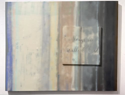 a horizontal rectangular canvas with vertical stripes of sprayed-on diffuse color in tan and blue shades in varied dimensions, the stripes transition from dark blue grey to light tan back to a near black, on top of the canvas on the right side is mounted another canvas, text peaks through the stripes of color in a readymade script printed on in a Michael's craft supply factory, reading in flowery calligraphy, "Always stay humble & kind."