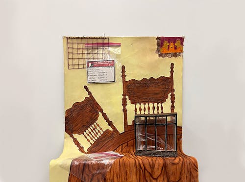 An oil painting with two wooden chairs and a wooden table. On the left side, there are two calendars stacked on top of one another made out of reddish-pink thread. There is also a ceramic insurance card in a ziplock bag that is pinned to the “wall” of the painting. On the right side, there is a painting of 3 chickens outlined in orange and a yellowish-light brown thread. The “table” of the painting is holding another ceramic insurance card with another ziplock bag. As well as a small building made out of ceramics.